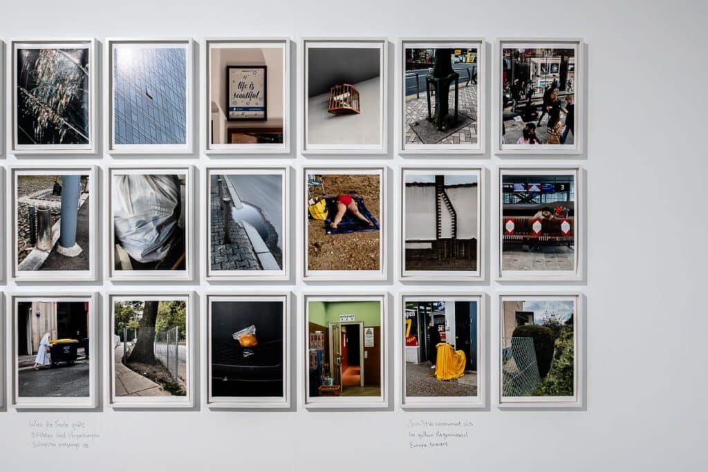 Ein Raster aus sechzehn gerahmten Fotografien, in der Ausstellung von Lars Eidinger, schmückt die weiße Wand. Jedes Foto fängt verschiedene urbane und natürliche Elemente ein. Unter den Rahmen sind handschriftliche Titel, die Haikus von Yoko Tawada sichtbar.