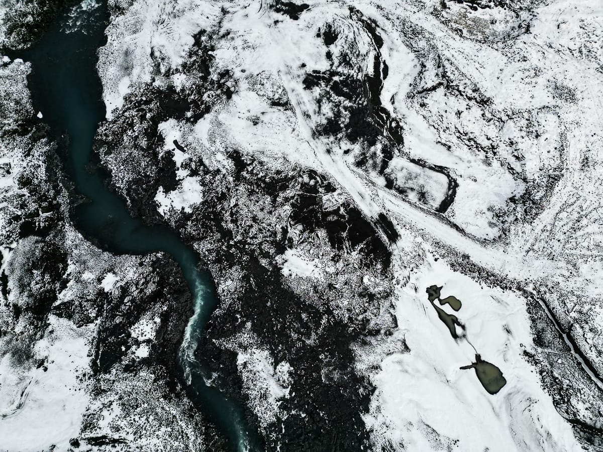 Luftaufnahme einer verschneiten Landschaft mit einem gewundenen Fluss und spärlicher Vegetation. Die Szene zeigt dunkle Felsformationen, die einen Kontrast zum Schnee bilden, und rechts zwei kleine zugefrorene Seen. Der Fluss schneidet durch das Gelände und erzeugt ein abstraktes natürliches Muster in der Winterlandschaft Islands.