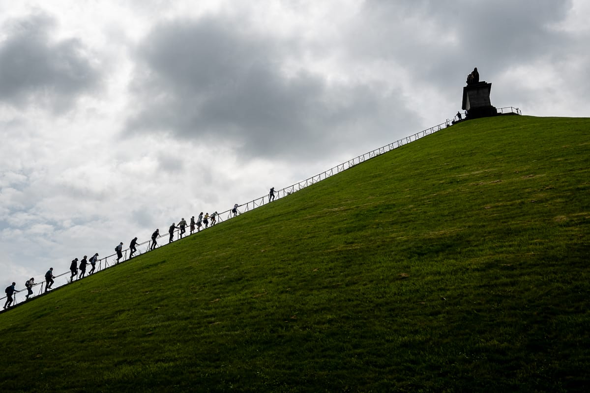 Eine Gruppe erklimmt unter einem wolkigen Himmel einen steilen, grasbewachsenen Hügel am Waterloo Schlachtfeld. Oben steht eine Löwenstatue auf einem Sockel. Der von einem Geländer gesäumte Hügel neigt sich steil von rechts nach links, während die langen Schatten der Menschen die Neigung und die bewölkte Beleuchtung betonen. Teil des Jahresrückblicks 2024.