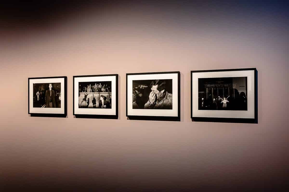 An einer Galeriewand hängen vier gerahmte Schwarzweißfotos. Jedes Bild zeigt eine andere Szene: Das erste zeigt eine stehende Person, das zweite eine versammelte Gruppe, das dritte zeigt eine Nahaufnahme einer Umarmung und das vierte zeigt eine Person in der Nähe eines Gebäudes, dem Brandenburger Tor.