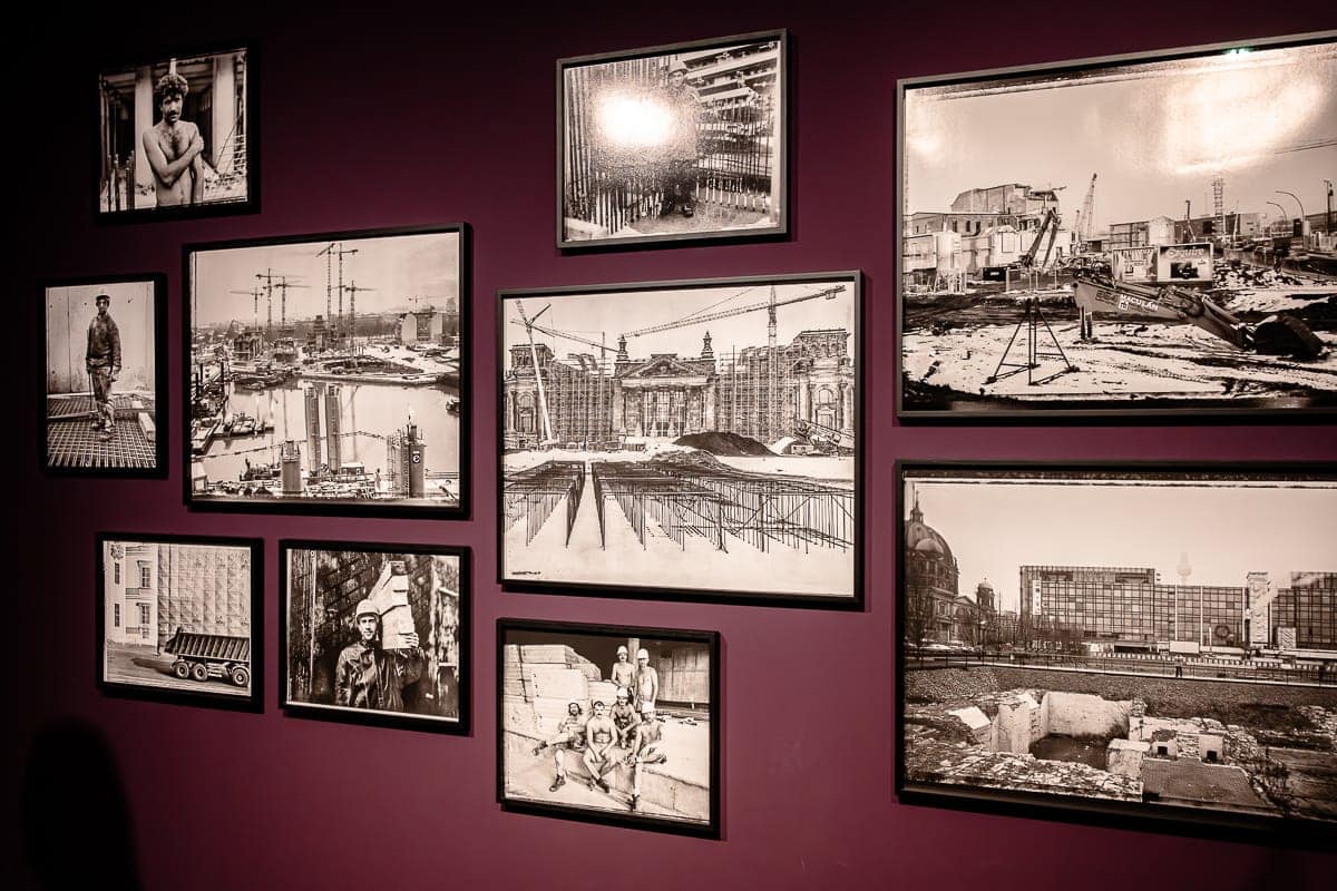 An einer Galeriewand hängen gerahmte Schwarzweißfotografien von Ostkreuz-Fotograf Maurice Weiss auf dunkelviolettem Hintergrund. Die Bilder zeigen verschiedene Stadtszenen, Baustellen und Porträts rund um den Potsdamer Platz. Die Anordnung ist asymmetrisch, mit unterschiedlichen Größen und Ausrichtungen, wodurch eine visuell dynamische Darstellung entsteht.
