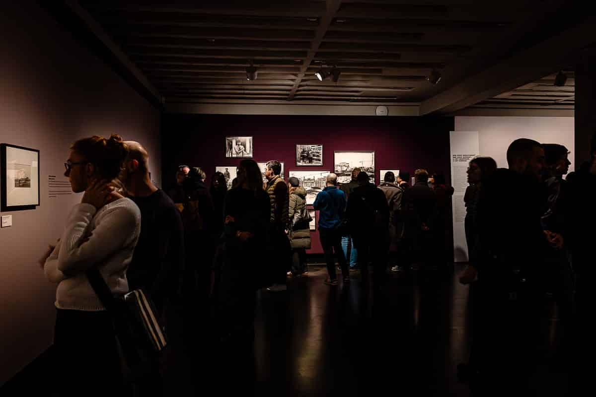 Menschen haben sich in einer schwach beleuchteten Kunstgalerie versammelt und betrachten gerahmte Fotografien der Fotoagentur Ostkreuz an den Wänden. Der Raum hat dunkelviolette Wände und Spotbeleuchtung. Besucher sind beschäftigt, einige stehen allein, andere in Gruppen. Die Decke hat ein Gittermuster mit Einbauleuchten.