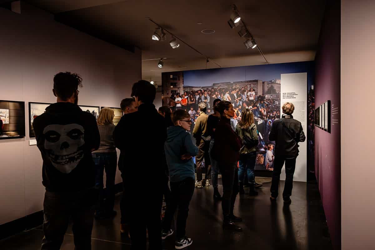 Eine Gruppe von Menschen betrachtet Kunst in einer schwach beleuchteten Galerie. Der Raum zeigt ein großes Foto an der Rückwand und kleinere gerahmte Bilder an der Seitenwand. Die Besucher sind leger gekleidet, einige tragen Jacken, und sie beschäftigen sich mit den Exponaten. Die Atmosphäre ist konzentriert und nachdenklich.