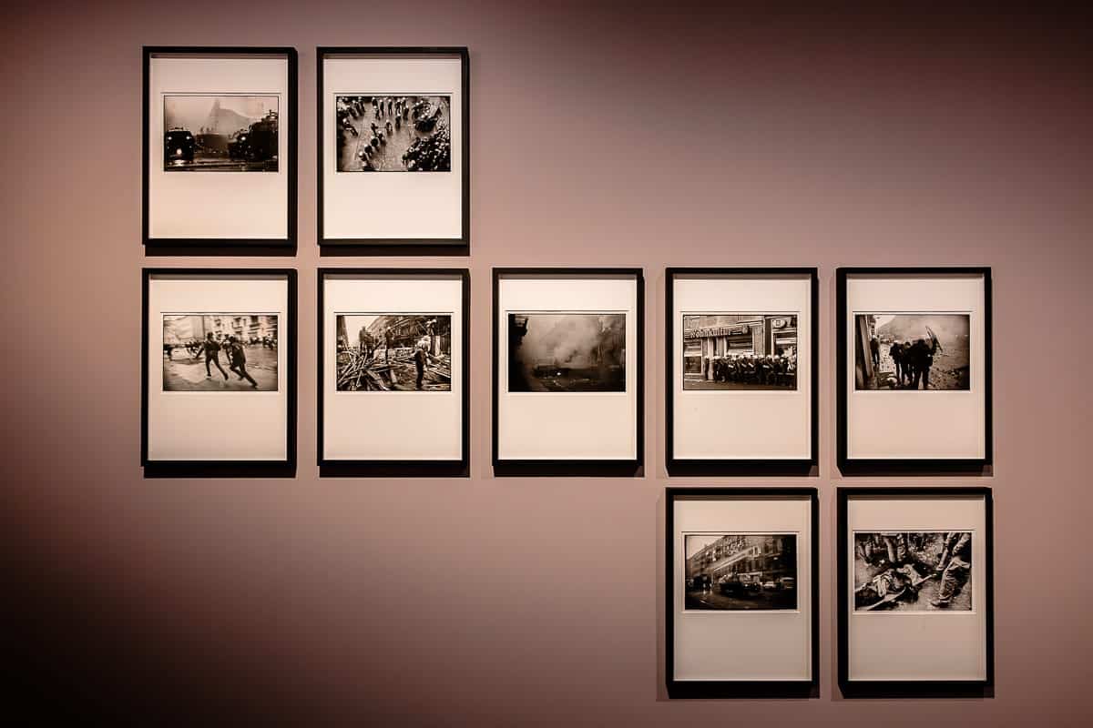 An einer Galeriewand hängen neun Schwarzweißfotos von Ostkreuz-Fotograf Harald Hauswald in schlichten Rahmen mit weißem Passepartout. Die Bilder zeigen verschiedene historische Szenen, angeordnet in drei Reihen mit einer ungleichmäßigen oberen und unteren Reihe. Die Wand ist sanft beleuchtet, was eine warme Atmosphäre schafft.