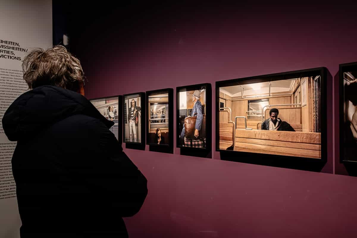 Eine Person mit kurzen, zerzausten Haaren und einer dunklen Jacke betrachtet gerahmte Fotografien an einer dunkelvioletten Wand in einer schwach beleuchteten Galerie. Die Kunstwerke zeigen verschiedene Szenen, darunter eine Person, die in einem leeren U-Bahn-Waggon sitzt, alles unter Glas mit schwarzen Rahmen.