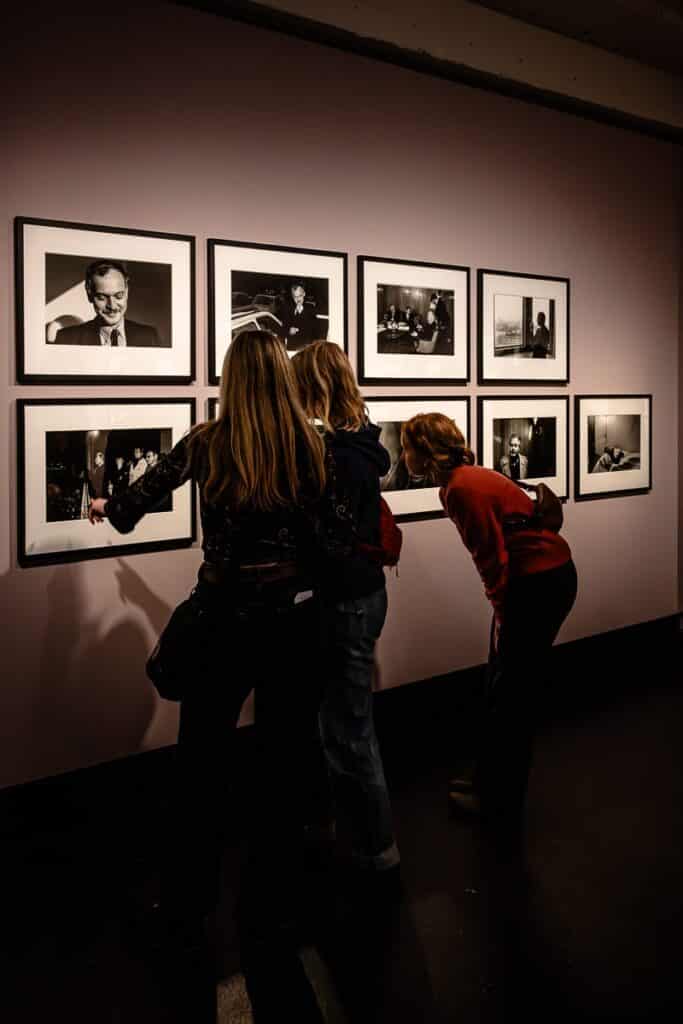 Drei Personen stehen da und betrachten eine Galeriewand im C/O Berlin mit neun gerahmten Schwarzweißfotos von Ostkreuz-Fotografin Ute Mahler. Der Raum ist schwach beleuchtet, wodurch die Bilder besonders gut zur Geltung kommen. Eine Person zeigt auf ein Foto, während sich andere interessiert vorbeugen. Die Wand ist in einem sanften Lila gehalten, das einen Kontrast zu den monochromen Bildern bildet.