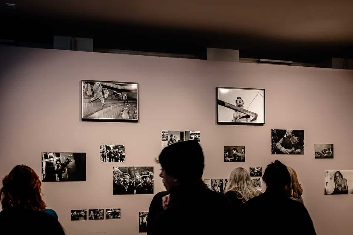 Menschen betrachten eine Galeriewand mit Schwarzweißfotos von Ostkreuz-Fotografin Ute Mahler, die verschiedene Motive und Szenen zeigen. Zwei größere gerahmte Bilder sind prominent über mehreren kleineren platziert. Die Betrachter, meist in dunkler Kleidung, heben sich teilweise als Silhouetten von der beleuchteten Wand ab.