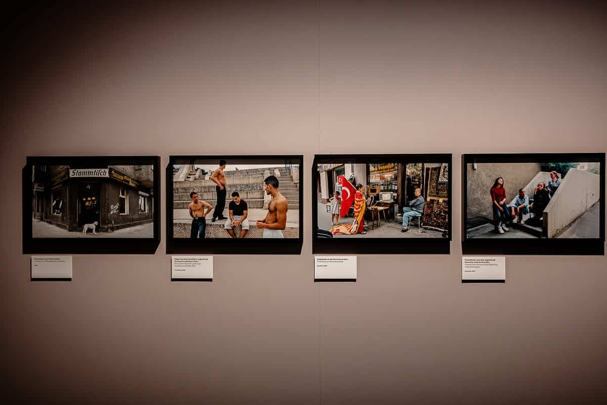 An einer Wand in einer Galerie hängen vier gerahmte Fotografien der Agentur Ostkreuz, darunter jeweils eine Schildchen mit einer Beschreibung. Die Fotos zeigen verschiedene Straßenszenen mit Menschen bei ihren täglichen Aktivitäten. Die Beleuchtung wirft einen warmen Schein auf die beige Wand und hebt die lebendigen Farben und Details in jedem Bild hervor.