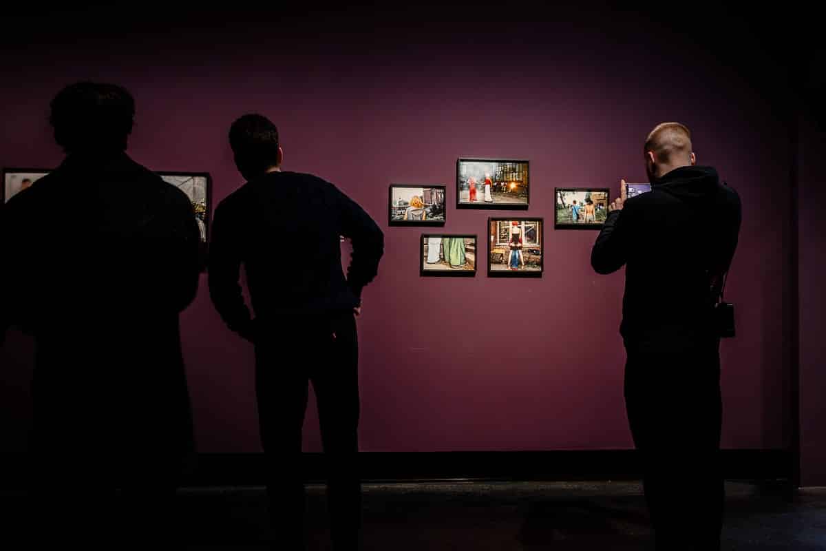 Drei Personen betrachten gerahmte Fotografien an einer dunkelvioletten Wand im C/O Berlin. Es sind neun Bilder der Agentur Ostkreuz, die asymmetrisch angeordnet sind. Die Beleuchtung erzeugt dramatische Schatten, die die kontemplative Atmosphäre verstärken. Die Gesichter der Personen sind nicht sichtbar, wodurch die Kunst und nicht die Betrachter im Mittelpunkt stehen.