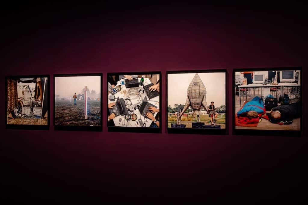 An einer dunklen Wand hängt eine Serie von fünf gerahmten Fotografien. Die Bilder zeigen verschiedene künstlerische Szenen: einen Mann mit einem glühenden Stab, eine Tischszene mit ungeordneten elektronischen Geräten und Händen, eine Gruppe um eine raketenförmige Struktur und Menschen in einem überfüllten Raum mit Computern und Sitzsäcken.