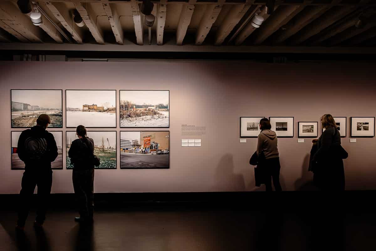 Menschen betrachten gerahmte Fotografien der Ostkreuz-Fotografen an einer schwach beleuchteten Galeriewand im C/O Berlin. Die Bilder scheinen städtische und ländliche Szenen darzustellen, sind aber im Mauerstreifen Berlin entstanden. Deckenleuchten sorgen für eine gezielte Beleuchtung. Besucher, die Winterkleidung tragen, stehen da und betrachten die Kunst, wodurch eine kontemplative Atmosphäre entsteht.