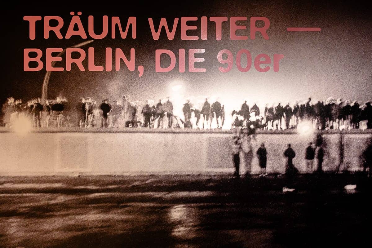 Eine Menschenmenge steht auf der Berliner Mauer, die sich in einer nächtlichen Szenerie vor hellen Lichtern abhebt. Der Text lautet in fetten Buchstaben „TRÄUM WEITER – BERLIN, DIE 90er“ und fängt die Atmosphäre Berlins in den 1990er Jahren ein. Das Bild hat ein nostalgisches, historisches Flair.