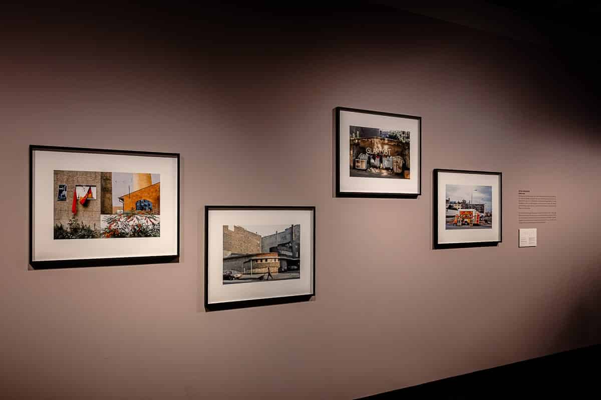 An einer dunklen Museumswand hängen vier gerahmte Fotografien von Ostkreuz-Fotografin Sibylle Bergemann. Jedes Foto zeigt eine urbane Szene mit unterschiedlichen Strukturen und Elementen des Stadtlebens. Rechts daneben informiert ein Wandtext über die Kunstwerke. Die Gesamtbeleuchtung hebt die Rahmen vor dem gedämpften Hintergrund hervor.