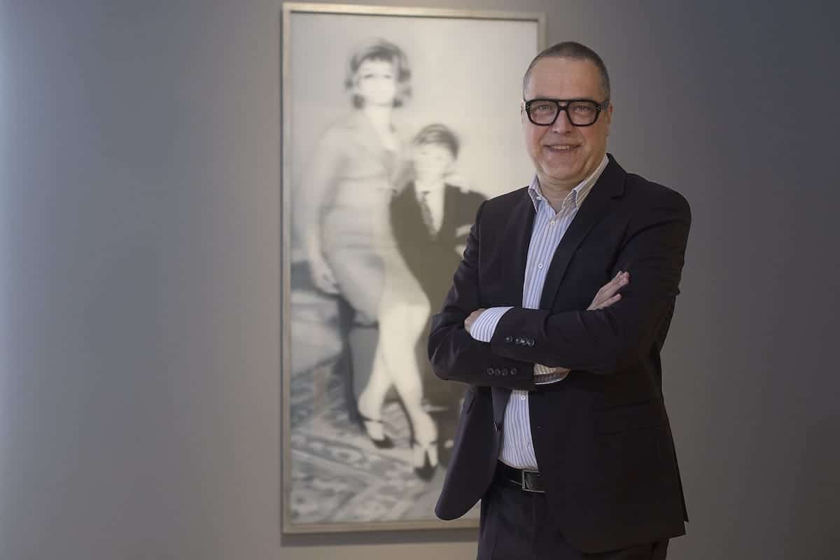 Ein Mann in schwarzem Anzug und Brille steht lächelnd mit verschränkten Armen vor einem großen Schwarzweißfoto. Das Foto zeigt eine sitzende Frau mit einem Kind neben ihr. Die Kulisse scheint eine Kunstgalerie mit grauen Wänden zu sein.
