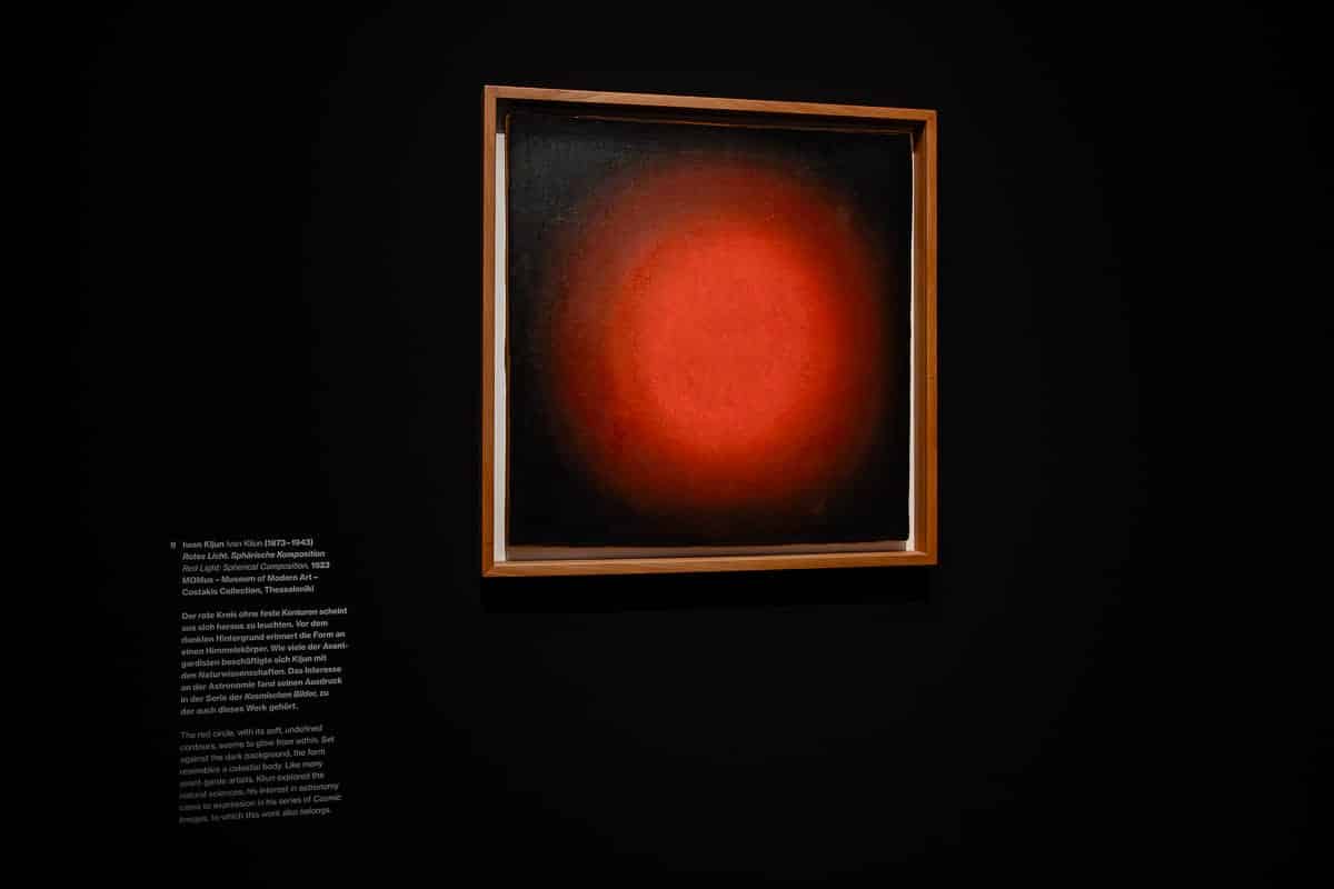Ein gerahmtes Gemälde einer leuchtend roten Kugel auf dunklem Hintergrund hängt an einer schwarzen Wand in einer Galerie. Links neben dem Gemälde befindet sich ein weißes Schild mit einer Beschreibung des Kunstwerks und des Künstlers. Die Beleuchtung hebt das lebendige, strahlende Zentrum hervor.