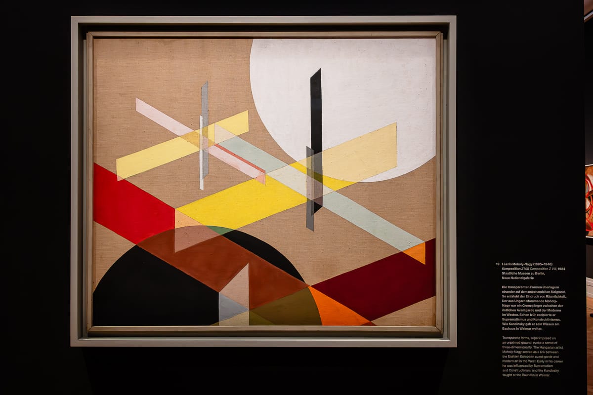 Ein gerahmtes abstraktes Gemälde zeigt sich kreuzende geometrische Formen: Kreise, Rechtecke und Linien in Weiß-, Rot-, Gelb-, Schwarz- und Brauntönen vor einem neutralen Hintergrund. Das Kunstwerk hängt an einer dunklen Wand in der Ausstellung Kosmos Kandinsky und trägt rechts ein Etikett mit einer Beschreibung.