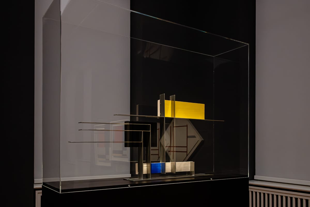 Eine moderne abstrakte Skulptur steht in einer Glasvitrine. Das Kunstwerk besteht aus geschichteten, rechteckigen Metall- und Farbtafeln in Schwarz, Silber, Gelb, Rot und Blau, die senkrecht zueinander angeordnet sind und so eine geometrische, minimalistische Komposition vor dunklem Hintergrund bilden.