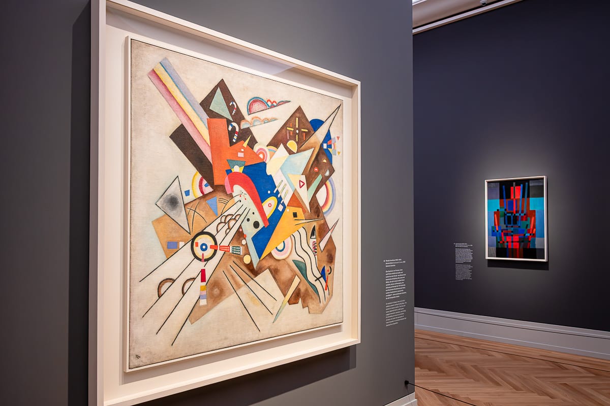 Eine Museumsgalerie mit grauen Wänden zeigt abstrakte Gemälde; im Vordergrund ist ein großes gerahmtes Kunstwerk von Wassily Kandinsky mit dynamischen geometrischen Formen und bunten Linien zu sehen, während an einer entfernten Wand ein kleineres, lebendiges Werk hängt. Der Boden besteht aus hellem Holz.