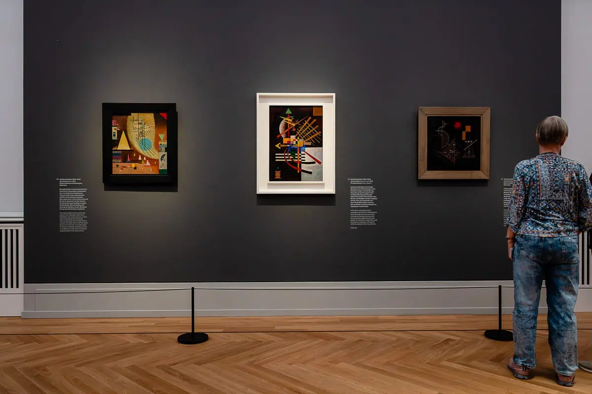 Eine Person in blau gemustertem Hemd und Jeans steht in einer Kunstgalerie vor einer dunkelgrauen Wand. Drei abstrakte Gemälde von Kandinsky sind gerahmt und im Scheinwerferlicht ausgestellt. Neben jedem Gemälde hängt ein Schild mit einer Beschreibung. Der Boden ist aus Parkett.