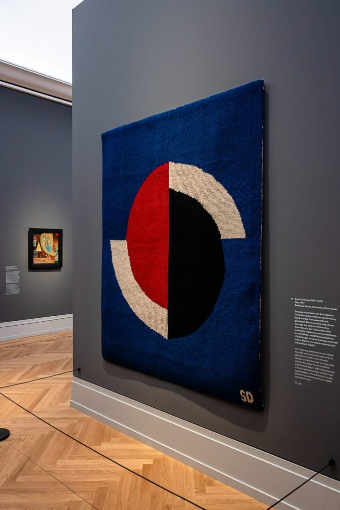 Ein geometrisches Textilkunstwerk mit kräftigen Formen – Halbkreisen in Rot, Schwarz und Weiß – auf tiefblauem Hintergrund hängt an der Wand einer Museumsgalerie. Die untere rechte Ecke ist mit „SD“ signiert. Ein kleineres gerahmtes Kunstwerk hängt an der angrenzenden Wand.