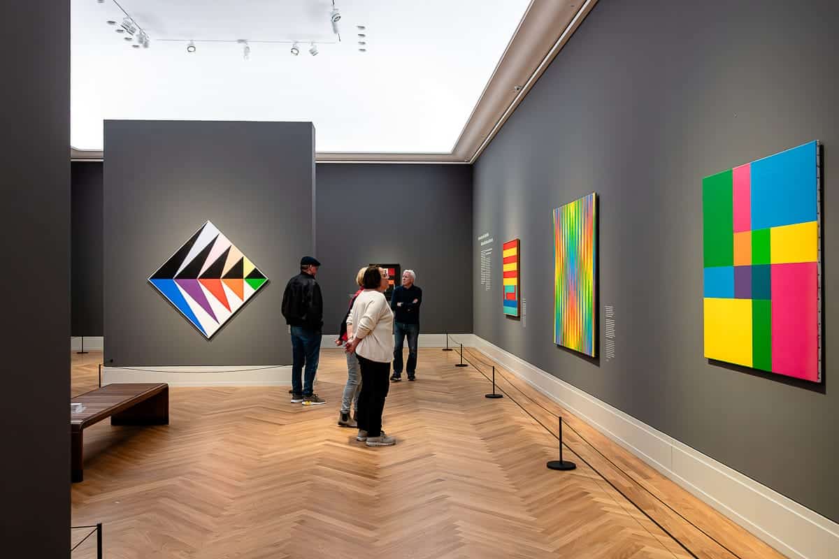 In einer modernen Kunstgalerie mit grauen Wänden und Holzböden können Besucher farbenfrohe abstrakte Gemälde mit kräftigen geometrischen Formen und leuchtenden Mustern betrachten. Vier Personen stehen und betrachten die Kunstwerke.