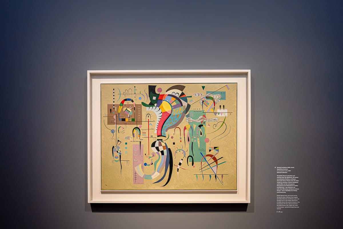 Ein farbenfrohes abstraktes Gemälde von Kandinsky in einem weißen Rahmen hängt an einer dunkelgrauen Wand. Das Kunstwerk zeichnet sich durch geometrische Formen, geschwungene Linien und kräftige Farben aus. Neben dem Gemälde informiert ein weißer Wandtext über das Werk und den Künstler.