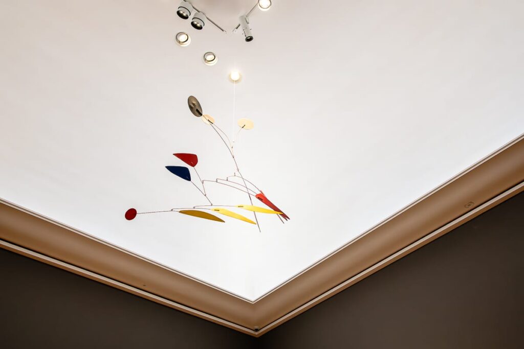 Decke mit Einbauleuchten und einem modernen Mobile mit abstrakten Metallformen in Rot, Gelb, Blau und Schwarz, die an dünnen Drähten hängen. Das Mobile wirft leichte Schatten auf die weiße Decke, die von einer beigefarbenen Zierleiste eingefasst ist.