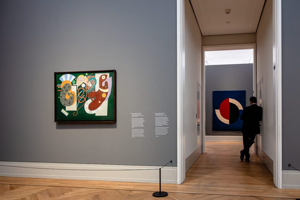 Eine moderne Kunstgalerie mit hellen Holzböden und grauen Wänden zeigt abstrakte Gemälde. Ein Gemälde mit leuchtenden Farben und Formen hängt links. Eine Person im dunklen Anzug geht durch eine Tür hinaus; im Nebenraum ist ein weiteres abstraktes Kunstwerk zu sehen.