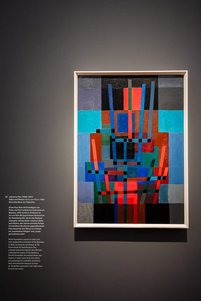 Ein gerahmtes abstraktes Gemälde mit überlappenden geometrischen Formen in Blau, Rot, Grün, Grau und Schwarz hängt an einer dunkelgrauen Wand. Links davon zeigt ein weißes Etikett den Namen des Künstlers und Details zum Kunstwerk in englischer Sprache. Das Werk ist hell beleuchtet, wodurch die leuchtenden Farben hervorgehoben werden.