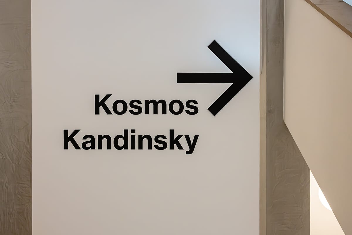Eine weiße Wand im Treppenhaus des Museum Barberini mit schwarzem Text trägt die Aufschrift „Kosmos Kandinsky“ und ein großer schwarzer, nach rechts zeigender Pfeil über den Worten weist die Richtung an. 