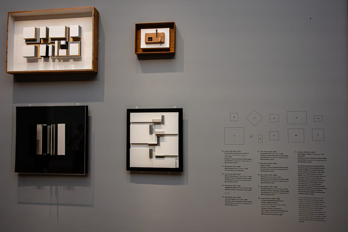 Vier geometrische 3D-Wandkunstwerke, hauptsächlich in Weiß-, Schwarz- und Holztönen, sind an einer grauen Wand angebracht. Rechts daneben befinden sich kleine Liniendiagramme und Textbeschriftungen, die jedes Kunstwerk beschreiben und in einem übersichtlichen Raster angeordnet sind.