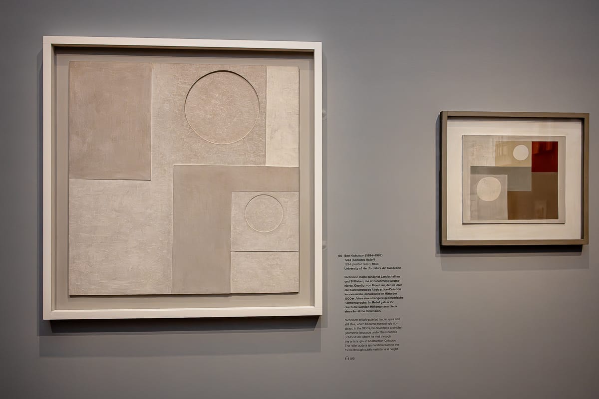 Zwei gerahmte abstrakte Kunstwerke hängen an einer grauen Wand. Beide Gemälde verwenden neutrale Töne und geometrische Formen, darunter Kreise und Rechtecke, die in strukturierten, minimalistischen Designs übereinandergelegt sind. Zwischen den beiden Werken befindet sich ein schwarzer Wandtextblock.