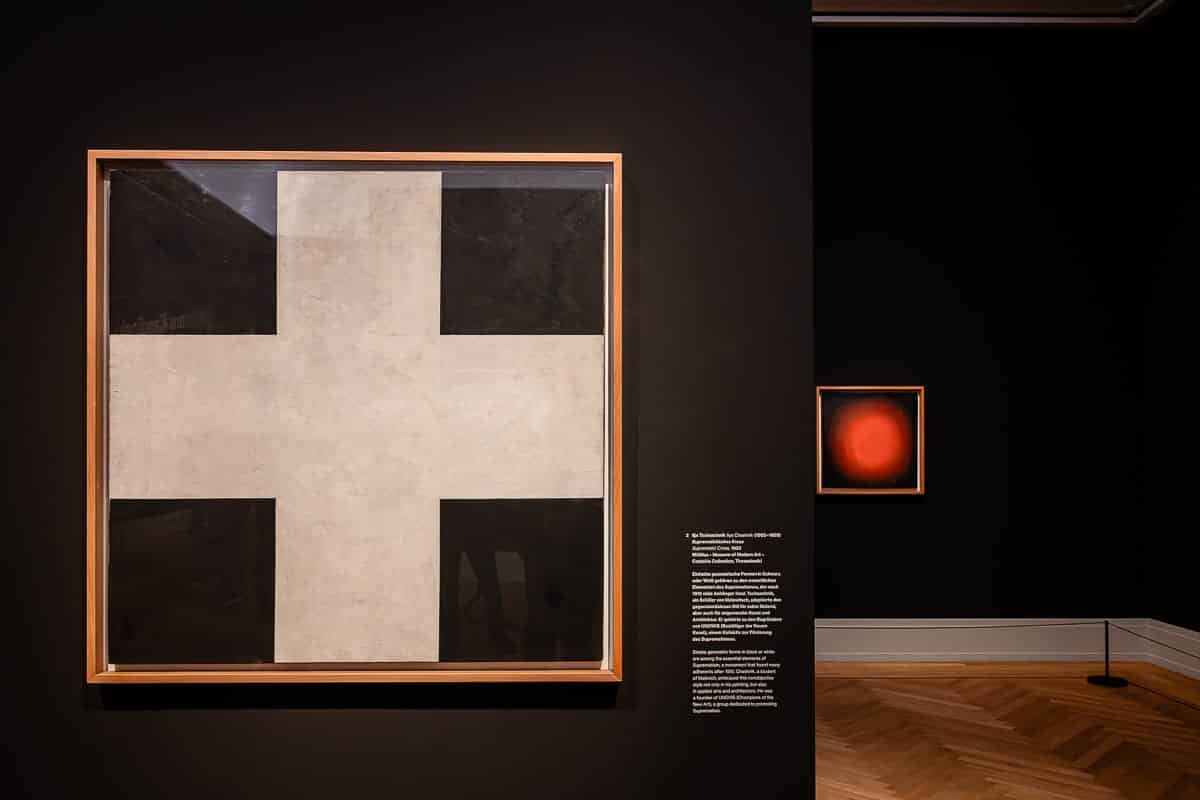 An einer dunklen Wand hängt ein gerahmtes abstraktes Gemälde mit einem großen weißen Kreuz auf schwarzem Hintergrund. Weiter hinten im Raum hängt ein kleineres gerahmtes Kunstwerk mit einer leuchtend roten Kreisform. Der Boden ist aus Holz, an der Wand ist ein Museumstext zu sehen.