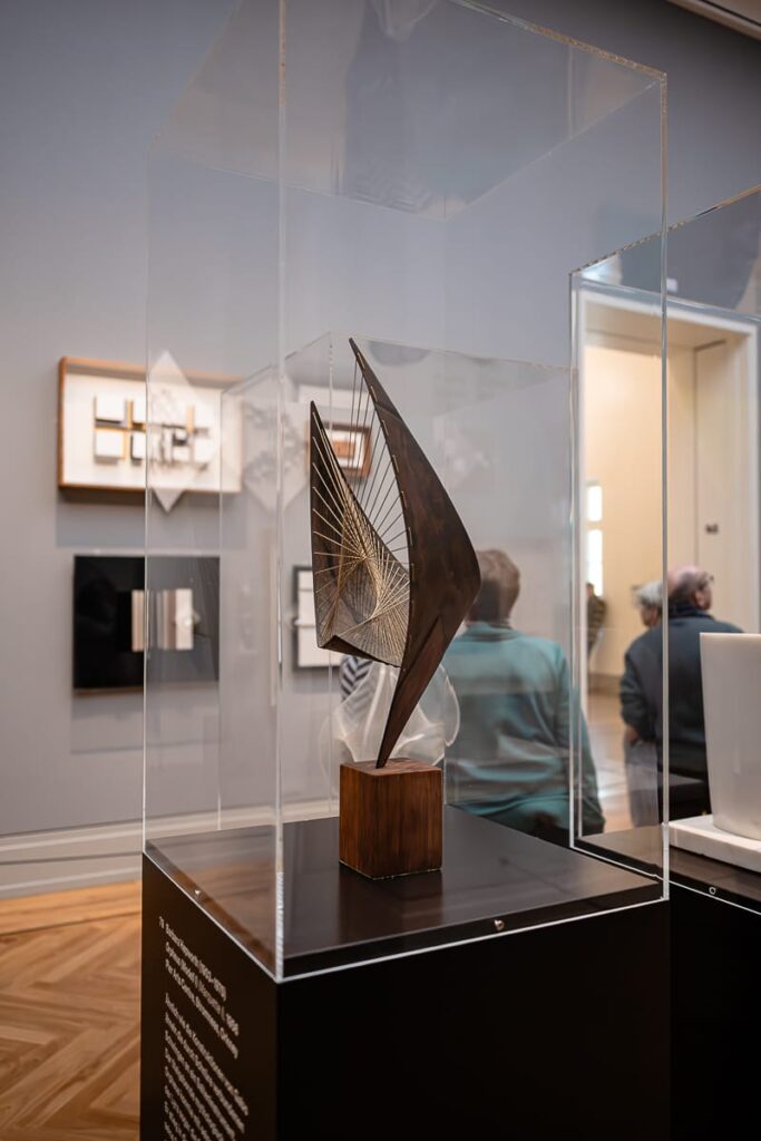 Eine moderne abstrakte Skulptur in einer durchsichtigen Glasvitrine auf einem Holzsockel. Die Skulptur besteht aus geschwungenen Metallformen und gespannten Schnüren, die geometrische Muster bilden. Im Hintergrund sitzen zwei Personen und betrachten Kunst an einer Museumswand mit gerahmten Werken.