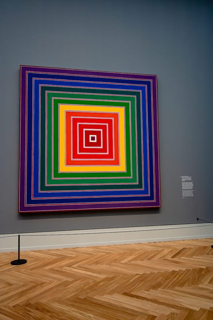 Ein großes quadratisches Kunstwerk mit konzentrischen Quadraten in leuchtenden Regenbogenfarben – Rot, Gelb, Grün, Blau und Lila – hängt an einer grauen Wand in einer Galerie mit Parkettboden. In der Nähe sind ein kleiner schwarzer Pfosten und eine Informationstafel zu sehen.