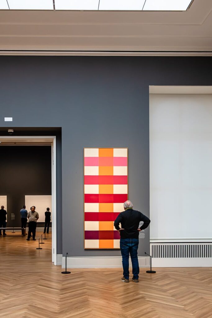 Ein Mann steht mit den Händen in den Hüften vor einem großen abstrakten Gemälde aus roten, rosa und orangefarbenen Quadraten an einer dunkelgrauen Wand in einer Kunstgalerie. Der Boden ist aus Holz, und durch eine Tür zu einem anderen Raum sind weitere Besucher zu sehen.