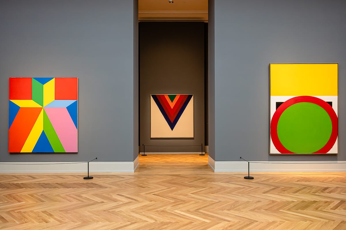 In einer Museumsgalerie mit hellen Holzböden und grauen Wänden sind drei abstrakte Gemälde ausgestellt: links ein geometrisches Werk mit sternförmigen Formen in leuchtenden Farben, in der Mitte eine Leinwand mit V-förmigen Streifen in Rot, Blau und Schwarz und rechts ein Gemälde mit einem großen grünen Kreis auf gelbem Hintergrund.
