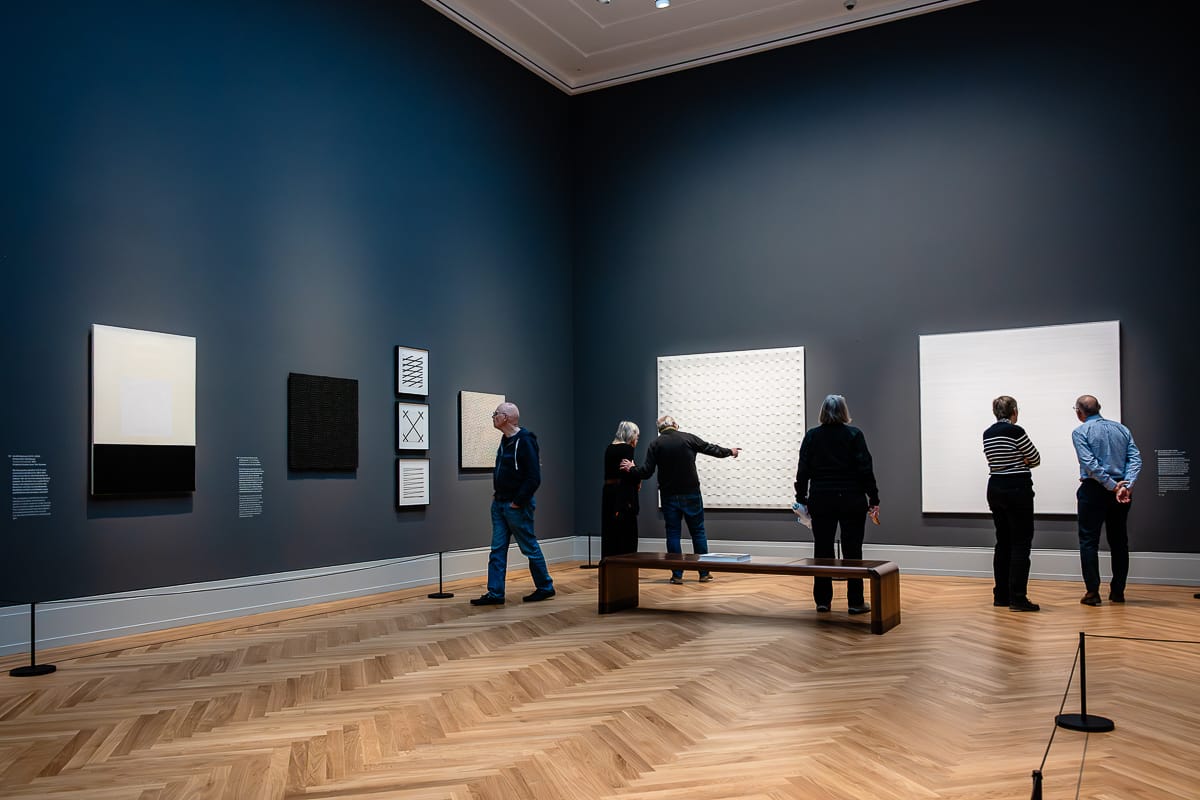 Fünf Personen betrachten minimalistische Kunst in der Ausstellung Kosmos Kandinsky an dunkelgrauen Wänden in einer geräumigen Galerie mit Fischgrätboden. Die Kunstwerke zeigen überwiegend schwarze, weiße und geometrische Muster. In der Mitte des Raumes steht eine Holzbank unter heller, gleichmäßiger Beleuchtung.