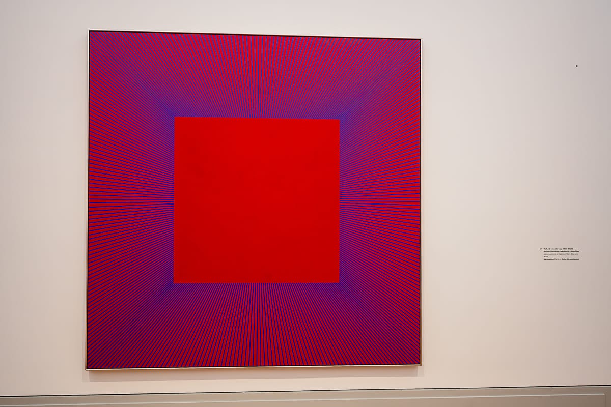 Ein großes, quadratisches abstraktes Kunstwerk mit leuchtend rotem Zentrum, umgeben von strahlenden blauen und magentafarbenen Linien, erzeugt einen optischen Täuschungseffekt in der Ausstellung Kosmos Kandinsky. Das Werk ist an einer weißen Galeriewand montiert, daneben befindet sich ein kleines Plakat.