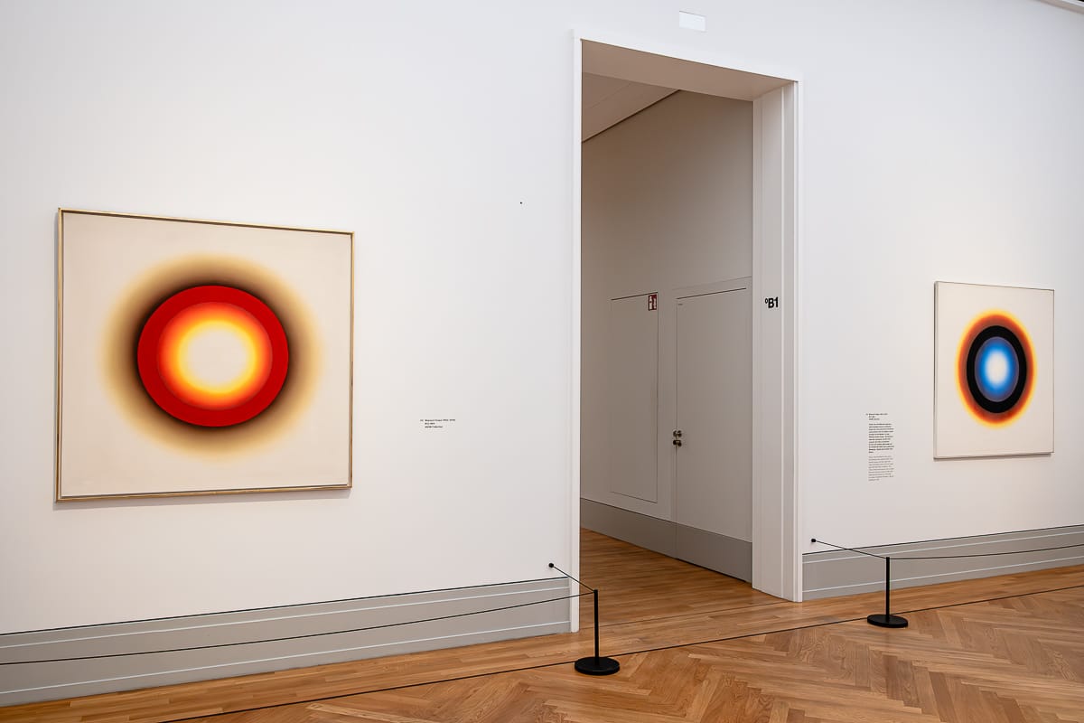 Eine moderne Kunstgalerie mit zwei abstrakten Gemälden an weißen Wänden. Das linke Gemälde zeigt einen rot-gelb leuchtenden Kreis, das rechte einen blau-orange leuchtenden Kreis. Der Raum hat Holzböden und einen Eingang im mittleren Hintergrund.