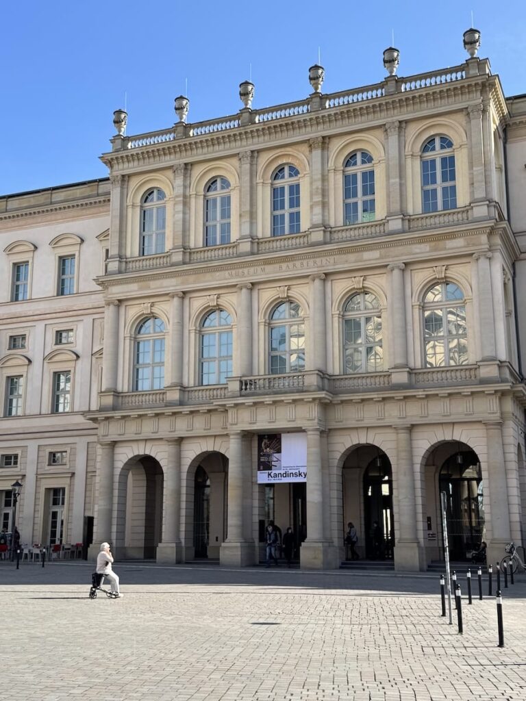Ein prächtiges neoklassizistisches Gebäude mit hohen Bogenfenstern und verzierten Säulen steht unter blauem Himmel. Auf einem Transparent steht „Kandinsky“. Eine Person mit einem Rollator geht über den gepflasterten Platz vor dem Gebäude.
