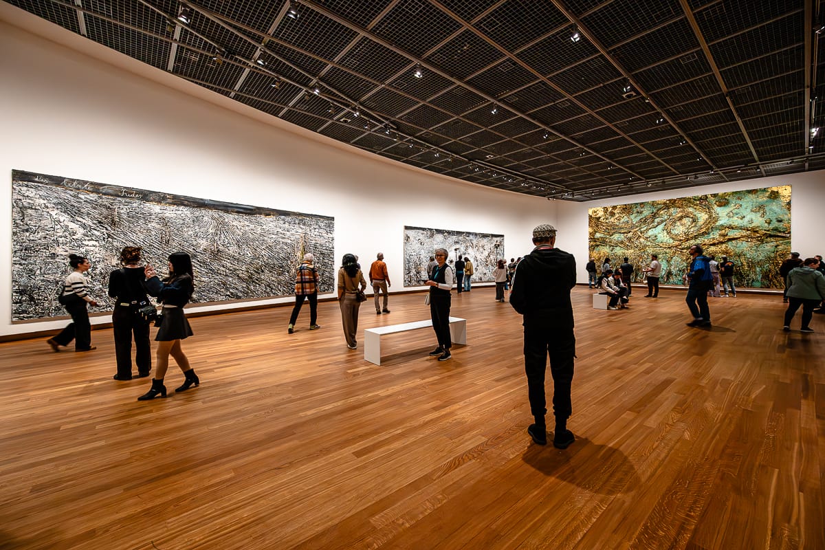 In einer geräumigen, modernen Kunstgalerie mit Holzboden, weißen Wänden und dunkler Rasterdecke werden große abstrakte Gemälde von Anselm Kiefer betrachtet. Besucher stehen, gehen und unterhalten sich, während sie die Kunstwerke betrachten, wodurch eine lebendige und engagierte Atmosphäre entsteht.