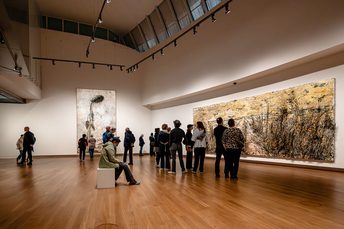 Eine Gruppe von Menschen betrachtet zwei große abstrakte Gemälde von Anselm Kiefer in einer geräumigen, modernen Kunstgalerie mit Holzboden und hohen Decken. Einige stehen vor dem Kunstwerk, eine Person sitzt auf einer weißen Bank. Alle sind in Ruhe mit den ausgestellten Werken beschäftigt.
