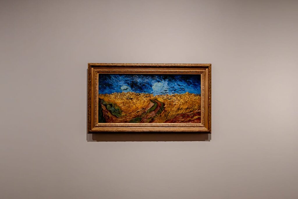 Ein gerahmtes Gemälde hängt an einer schlichten beigefarbenen Wand. Es zeigt ein Weizenfeld unter einem strahlend blauen Himmel mit wirbelnden Wolken und einem geschwungenen Pfad durch goldgelben Weizen und grüne Pflanzen. Der Malstil wirkt ausdrucksstark und ausdrucksstark.
