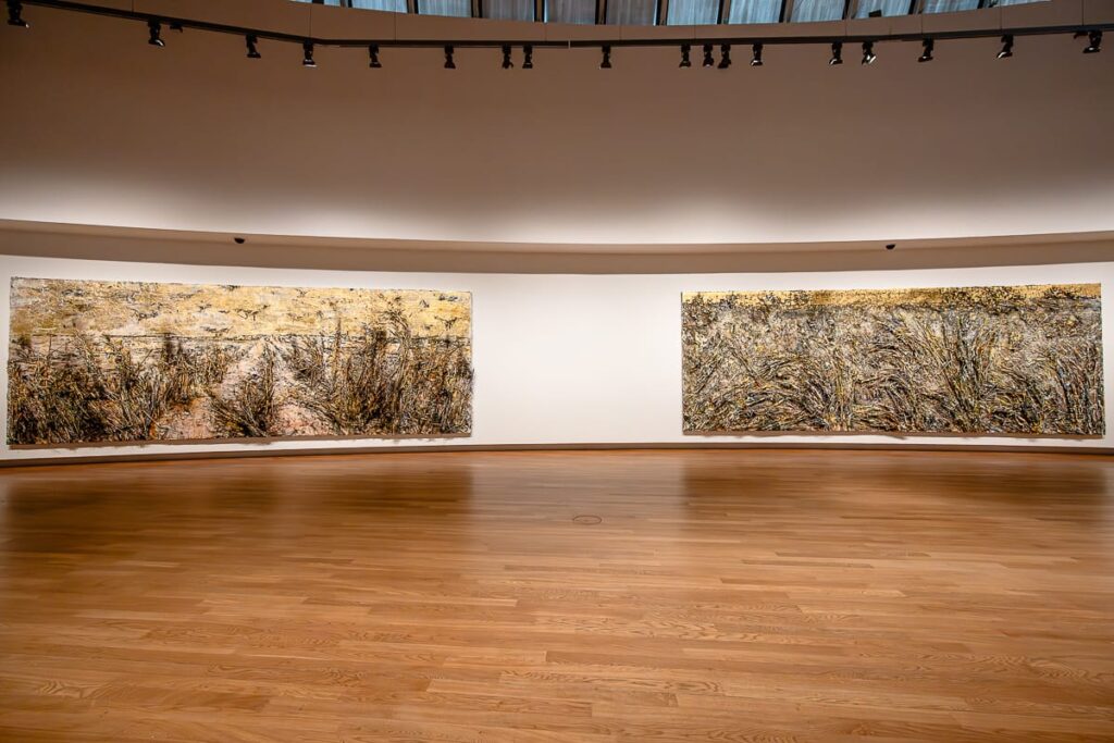 Eine geräumige Galerie mit Holzboden und einer geschwungenen weißen Wand zeigt zwei große abstrakte Landschaftsgemälde in Erdtönen von Anselm Kiefer, strukturierten Pinselstrichen und laubähnlichen Mustern. Scheinwerfer beleuchten das Kunstwerk von oben.