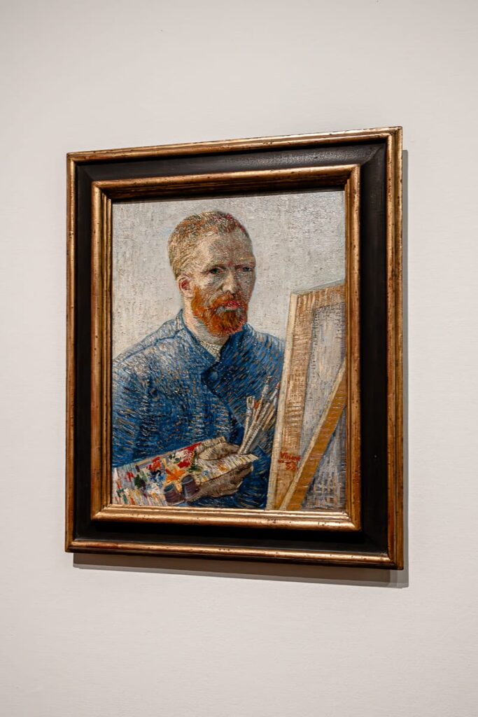 Ein gerahmtes Gemälde an einer weißen Wand zeigt einen bärtigen Mann mit rötlichem Haar in einem blauen Mantel - Van Gogh. Er hält Pinsel und Palette in der Hand und malt an einer Staffelei. Das Kunstwerk verwendet kräftige Pinselstriche und helle Farben und erinnert an einen impressionistischen Stil.