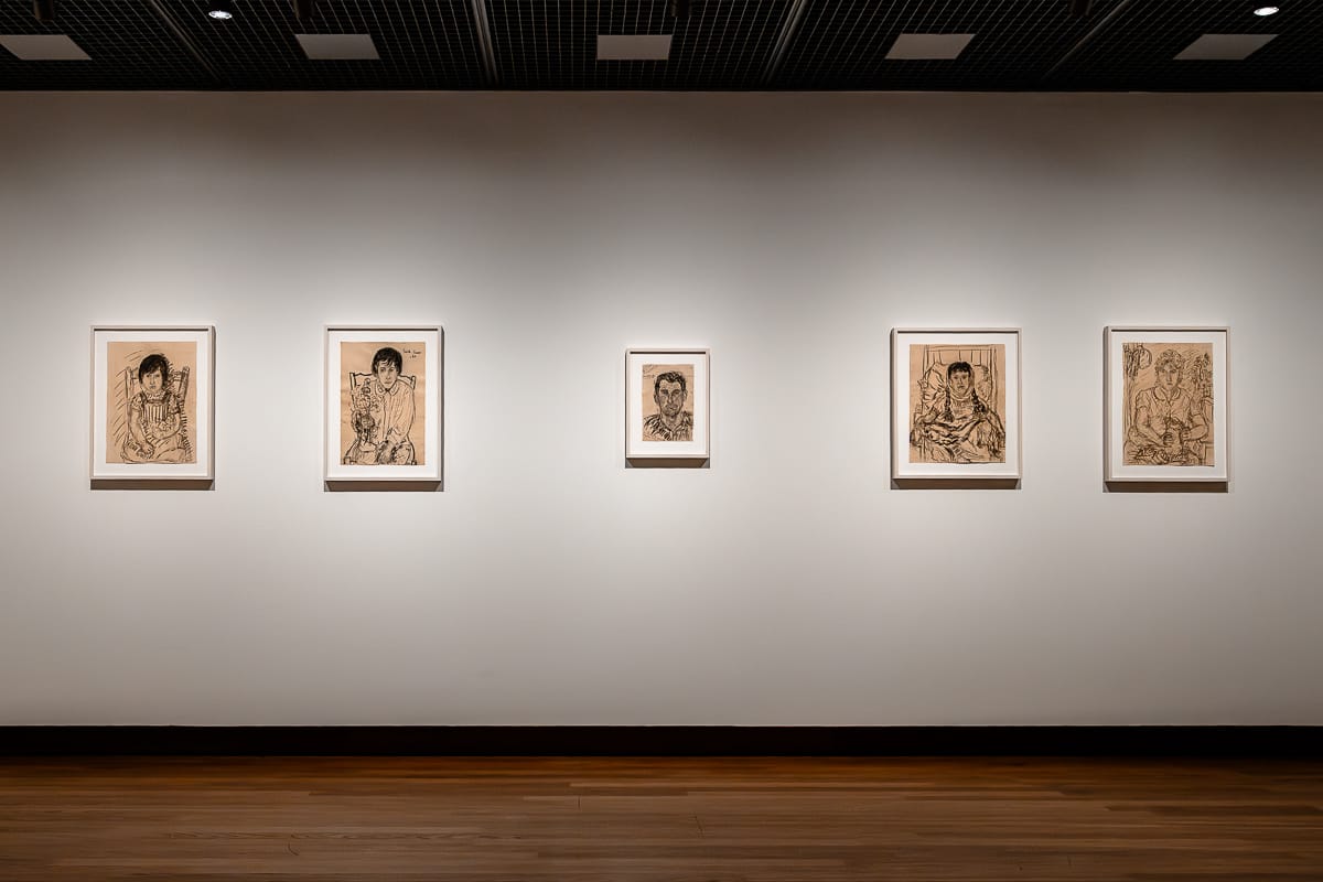 An einer Galeriewand hängen fünf gerahmte Schwarz-Weiß-Porträtzeichnungen von Anselm Kiefer in gleichmäßigem Abstand. Die Porträts zeigen einzelne Personen in detailreicher, ausdrucksstarker Strichzeichnung. Der Galerieraum ist mit Holzböden, weißen Wänden und einer dunklen Gitterdecke ausgestattet.