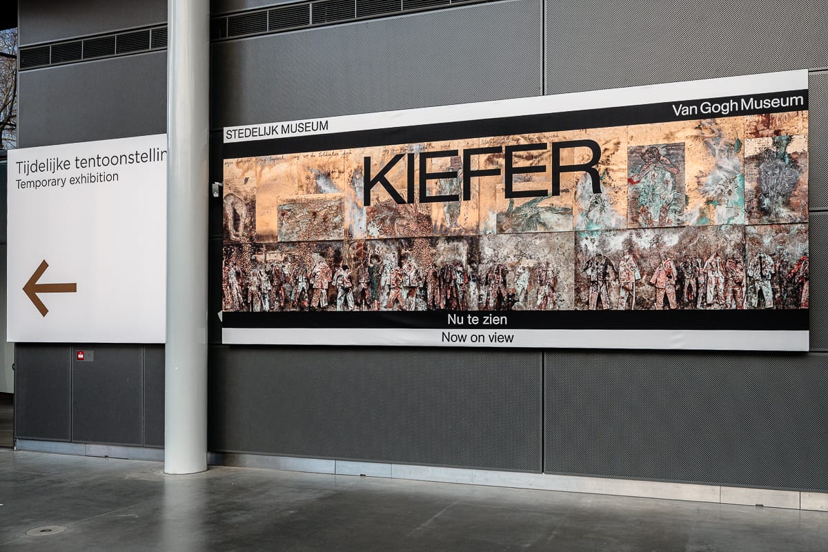 Ein großes Wandplakat im Van Gogh Museum Amsterdam wirbt für die Ausstellung „KIEFER“ mit einem vielschichtigen, strukturierten Kunstwerk. Auf dem daneben stehenden Schild steht auf Niederländisch und Englisch „Temporäre Ausstellung“ mit einem nach links zeigenden Pfeil. Die Ausstellung ist modern und in Innenräumen gestaltet.