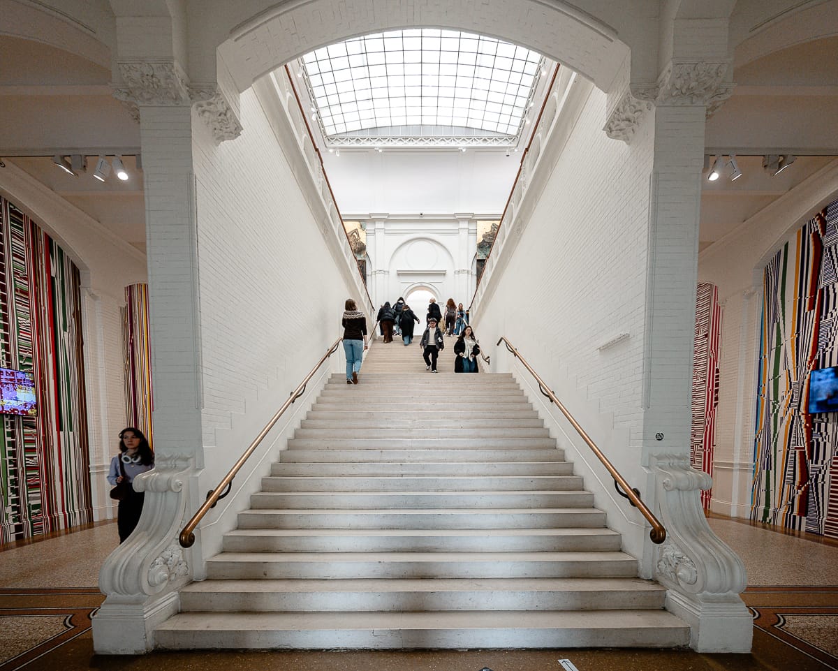Eine Gruppe von Menschen geht in einem prachtvollen Gebäude mit hohen Decken und einem Glasdach eine breite, weiße Steintreppe auf und ab. Verzierte Details schmücken die Treppe, und bunte vertikale Streifen zieren die Wände auf beiden Seiten.