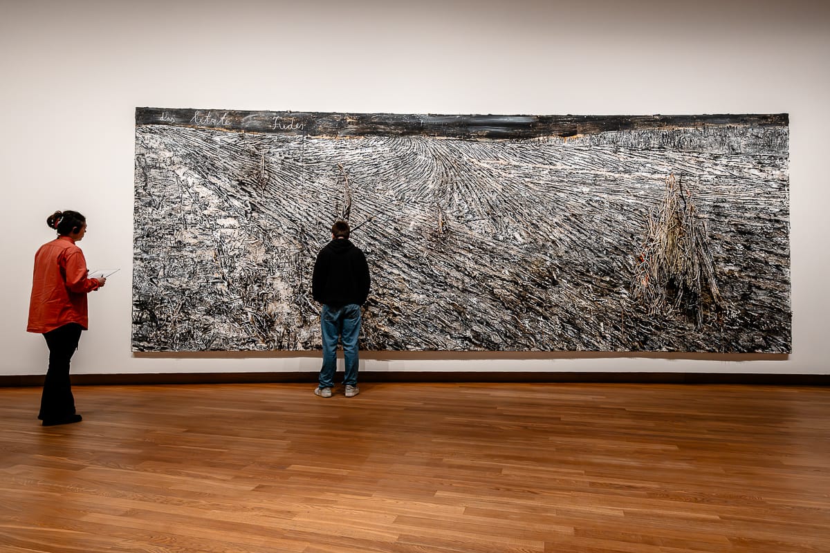Zwei Personen stehen in einer Galerie vor einem großen, strukturierten Landschaftsgemälde in neutralen Tönen von Anselm Kiefer. Eine Person trägt ein rotes Hemd und hält ein Notizbuch, während die andere, dunkel gekleidet, näher am Kunstwerk steht. Die Galerie hat Holzböden und schlichte Wände.